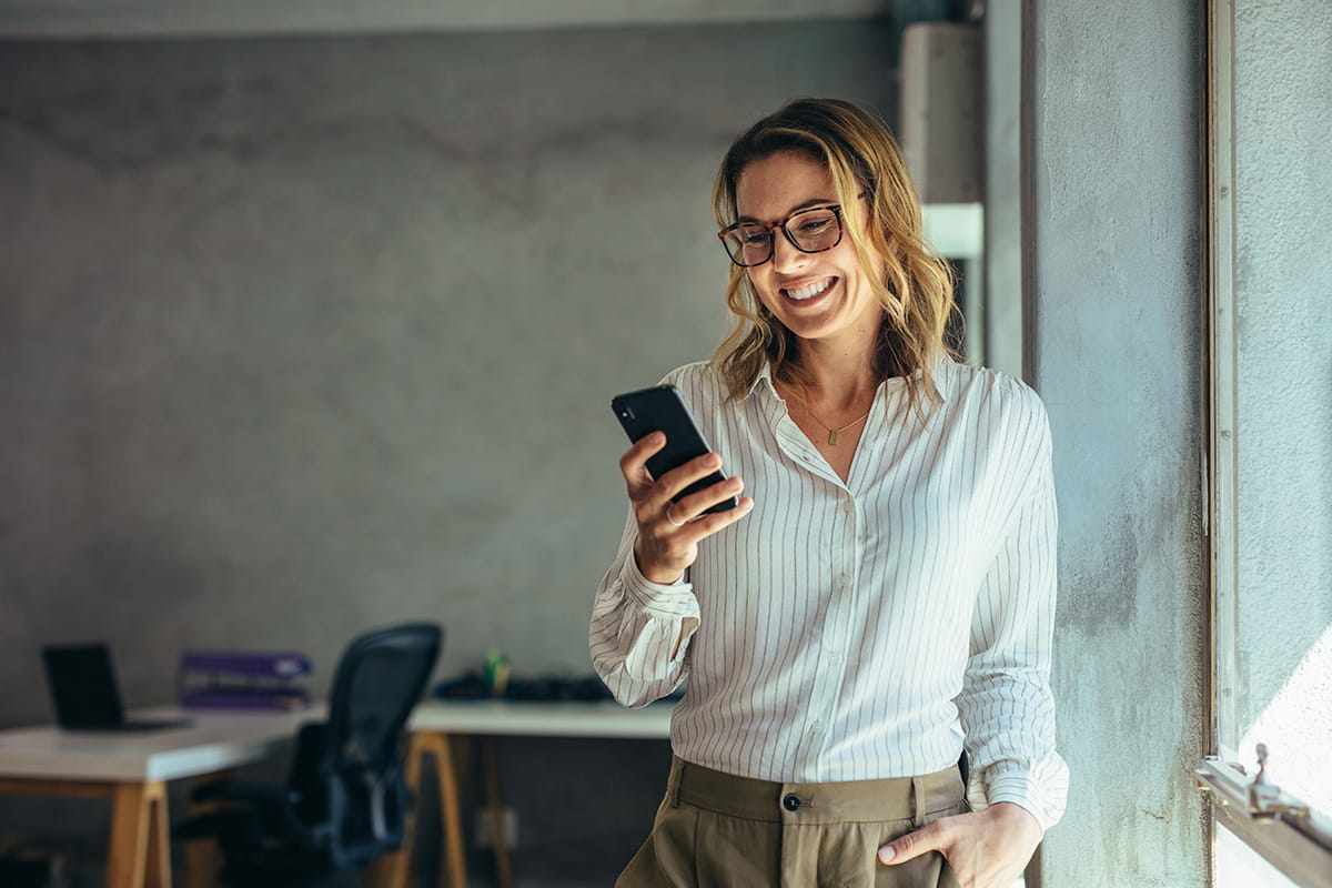 En smilende kvinde står ved et vindue og kigger på sin mobiltelefon i et moderne kontormiljø.