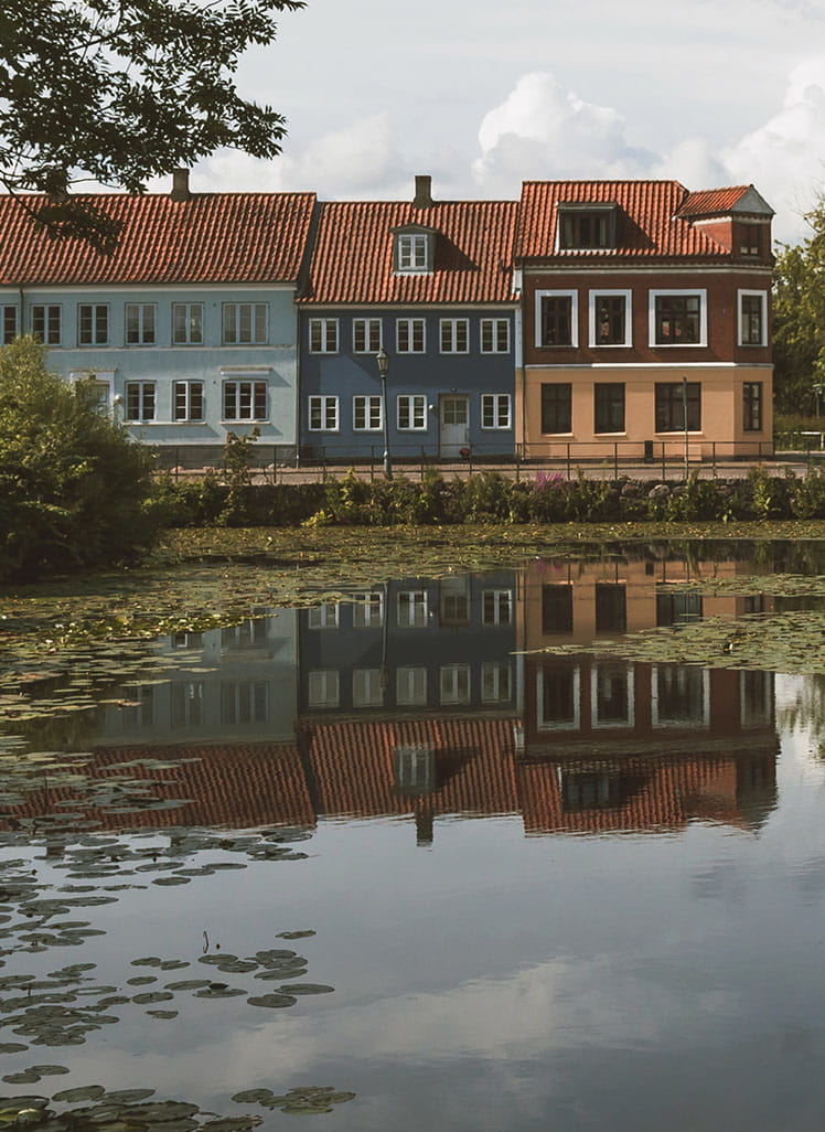 Boliger med røde tegltage set fra den anden side af en sø, omgivet af grønne træer og spejlinger i vandet på en rolig dag