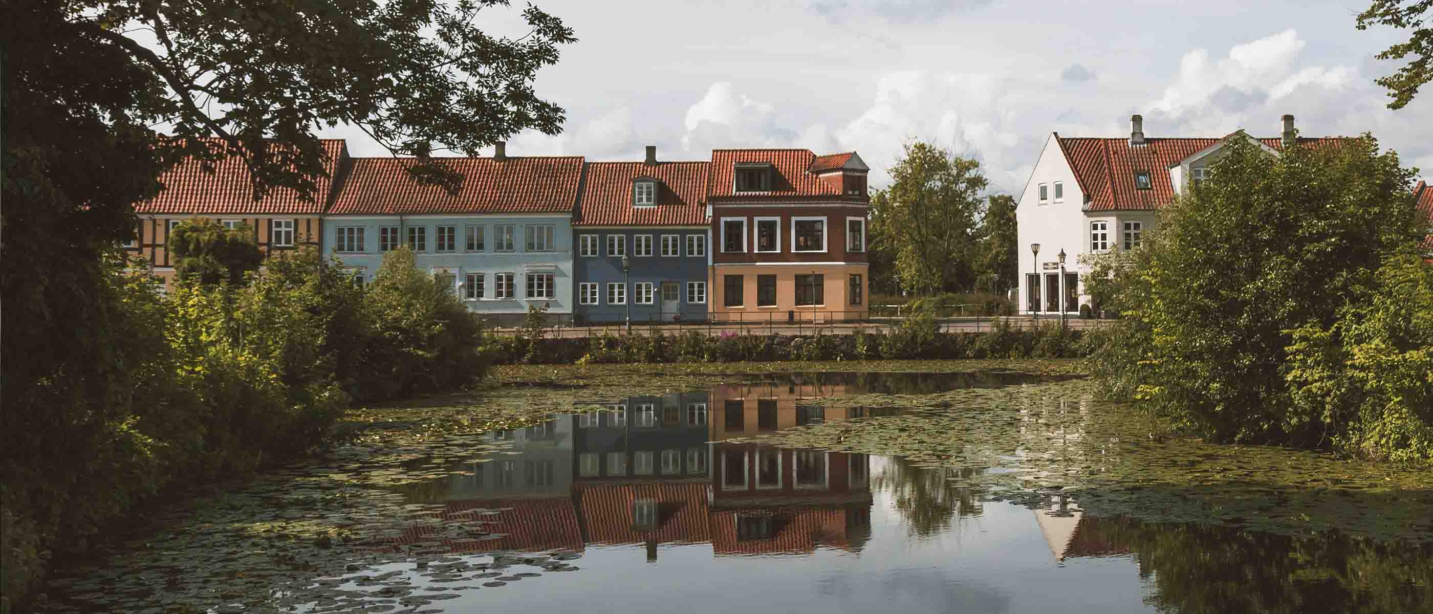 Boliger med røde tegltage set fra den anden side af en sø, omgivet af grønne træer og spejlinger i vandet på en rolig dag