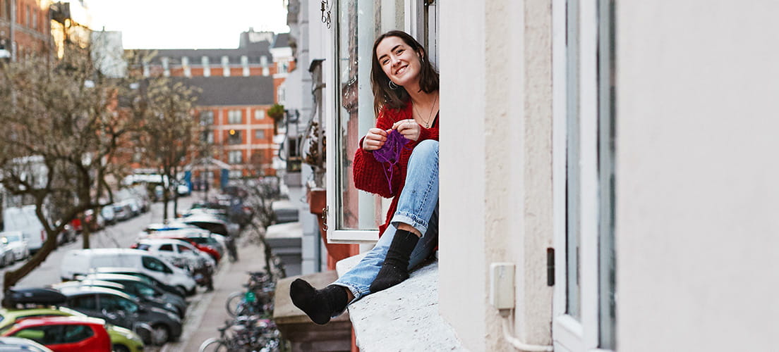 Kvinde sidder i et åbent vindue i en lejlighed og strikker, mens hun smiler ud mod gaden med parkerede biler og et bykvarter i baggrunden