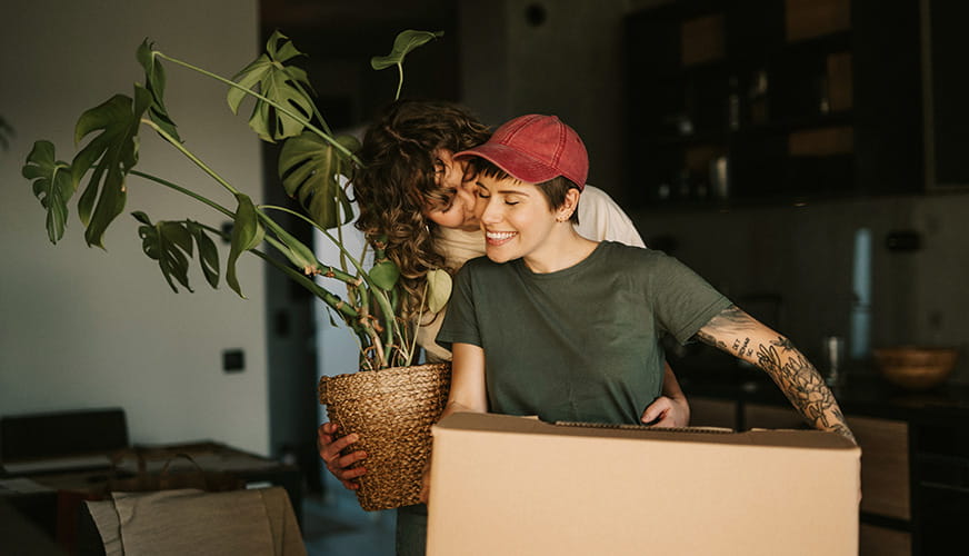 Par står tæt i deres nye hjem under indflytning, mens den ene holder en flyttekasse og den anden bærer en stueplante