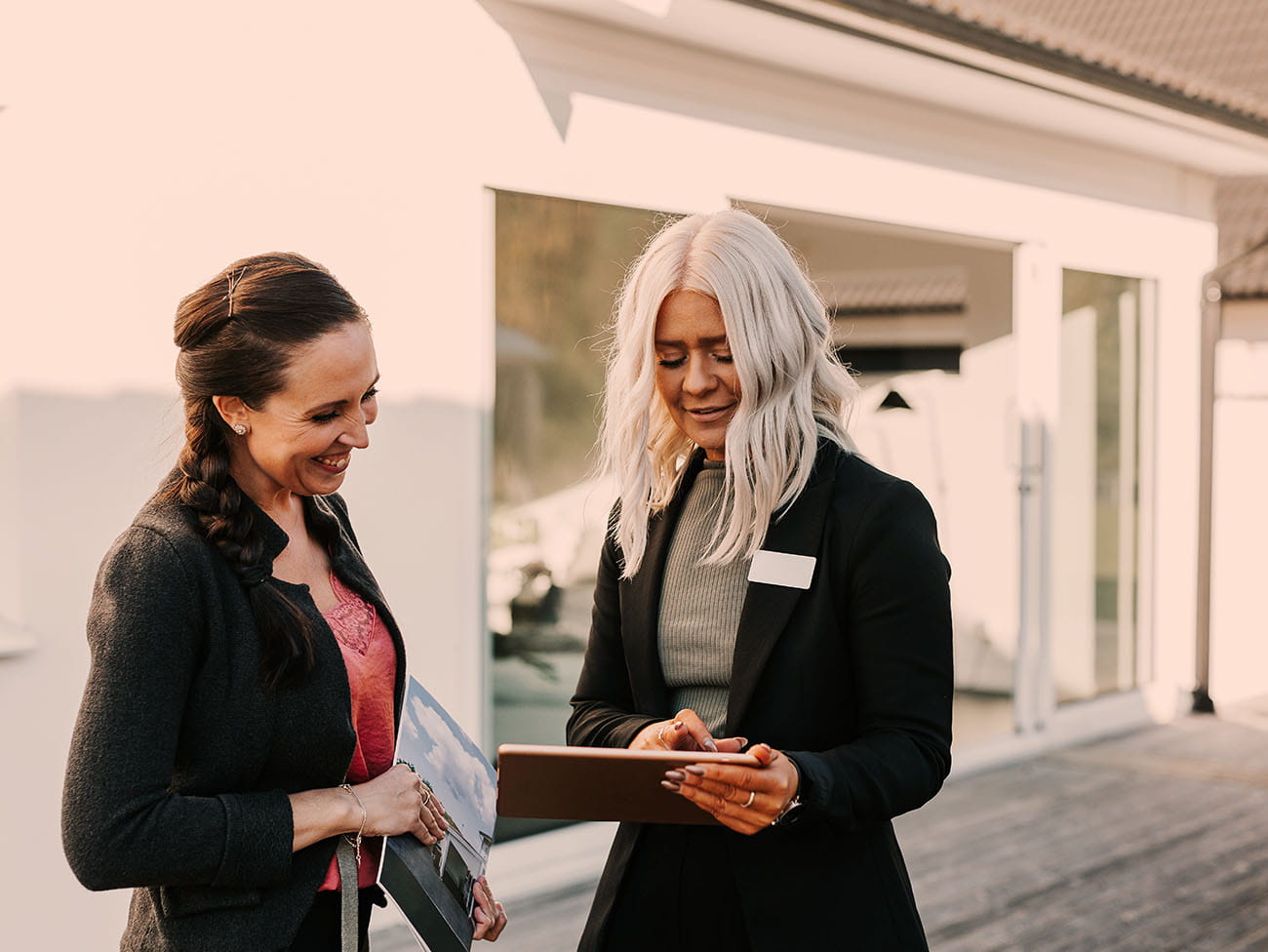 Boligrådgiver viser information på tablet til kvinde udenfor moderne bolig med terrasse og store vinduer
