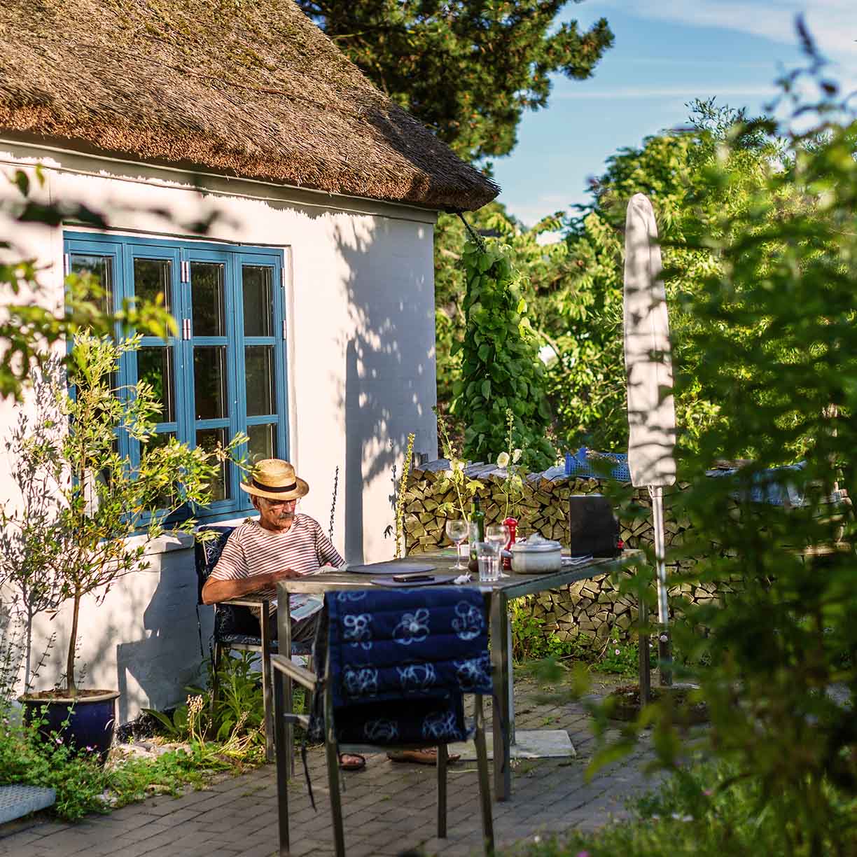 Mand sidder ved havebord foran sommerhus med stråtag og blå vinduer omgivet af grøn have