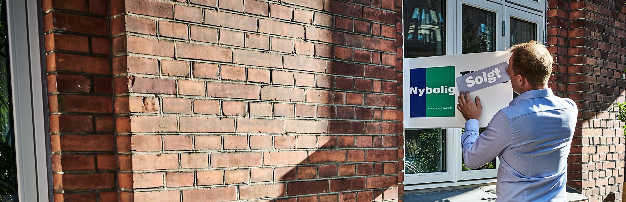 En mægler sætter et Nybolig Solgt-skilt på vinduet i en murstensbygning, der indikerer, at en fast ejendom er solgt